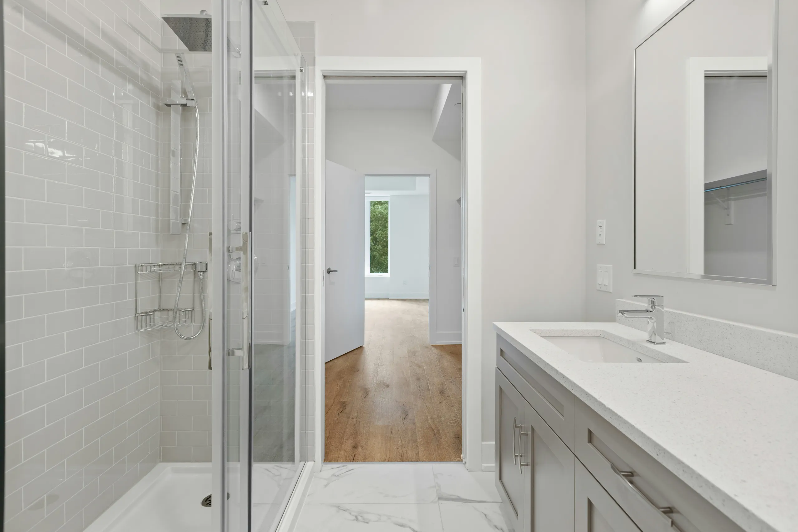 Baño luminoso con azulejo blanco, ducha tras cristal y vistas al dormitorio con suelo de madera